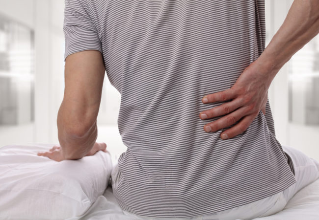 Man suffering from back pain at home in the bedroom. Uncomfortable mattress and pillow causes back pain.