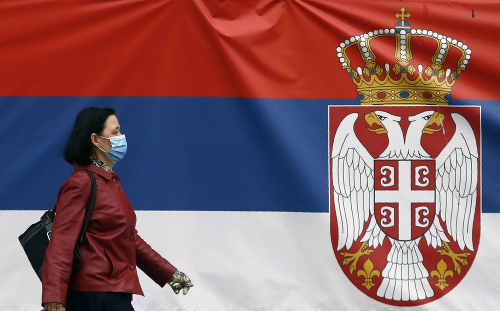 A woman wearing a face mask walks by a Serbian flag placed on a street prior a curfew set up to help prevent the spread of the new coronavirus in Belgrade, Serbia, Monday, April 13, 2020 (AP Photo/Darko Vojinovic)