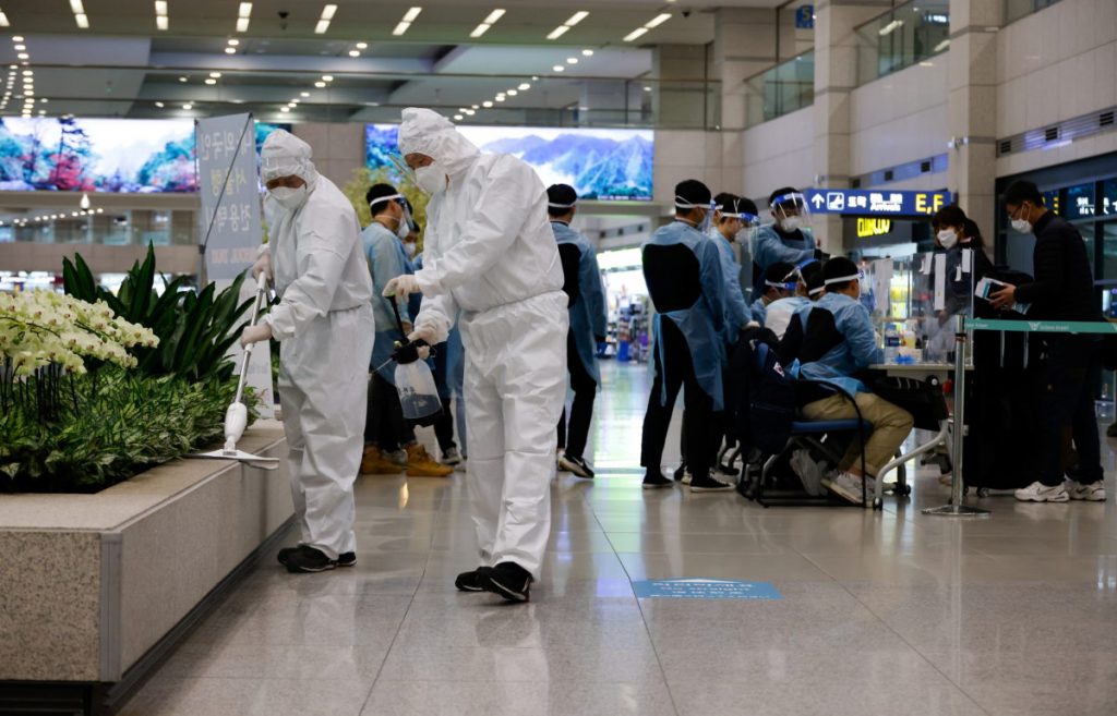 Workers wearing protective gear disinfect an arrival gate as other workers check passengers from overseas upon their arrival at the Incheon International Airport, in Incheon