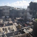 Residents stand amid their homes damaged by a gasoline truck that overturned and exploded in Cap-Haitien, Haiti, Tuesday, Dec. 14, 2021. The explosion engulfed cars and homes in flames, killing more than 50 people and injuring dozens of others. (AP Photo/Joseph Odelyn)