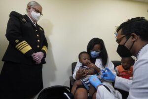 Pediatrician Emy Jean-Marie, center, holds her nine-month-old son Adedeji Adebayo, Emiola Adebayo, 3, on her lap as Dr. Nizar Dowla, right, administers a vaccine while        Department of Health and Human Services Assistant Secretary for Health, Admiral Rachel Levine, left, looks on, Tuesday, June 28, 2022, at the Borinquen Health Care Center in Miami. Florida is the only state that didn’t pre-order the under-5 vaccine, and state Surgeon General Joseph Ladapo has recommended against vaccinating healthy children. (AP Photo/Wilfredo Lee)