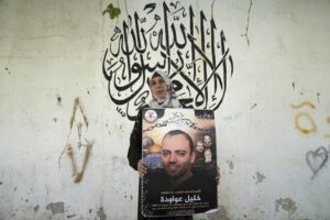 Palestinian Dalal Awawdeh poses with a poster of her husband Khalil Awawdeh, who is a prisoner in Israel, at the family house, in the West Bank village of Idna, Hebron, Tuesday, Aug. 9, 2022. Awawdeh, who is on a protracted hunger strike, was moved Thursday from an Israeli jail to a hospital because of his worsening condition, the prisoner’s wife said. Arabic in the background reads “There is no God but Allah, Muhammad is the Messenger of Allah.” (AP Photo/Nasser Nasser)