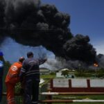 Workers of the Cuba Oil Union, known by the Spanish acronym CUPET, watch a huge rising plume of smoke from the Matanzas Supertanker Base, as firefighters work to quell a blaze which began during a thunderstorm the night before, in Matazanas, Cuba, Saturday, Aug. 6, 2022. Cuban authorities say lightning struck a crude oil storage tank at the base, causing a fire that led to four explosions which injured more than 50 people. (AP Photo/Ramon Espinosa)