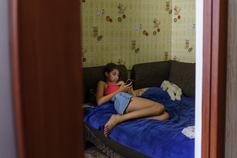 Anastasiia Aleksandrova, 12, looks at her phone in her bedroom in Sloviansk, Donetsk region, eastern Ukraine, Monday, Aug. 8, 2022. Anastasiia’s retreat into digital technology to cope with the isolation and stress of the war that rages on the front line only 7 miles (12 kilometers) away is an increasingly common phenomenon among young people in Ukraine’s embattled Donetsk region. (AP Photo/David Goldman)
