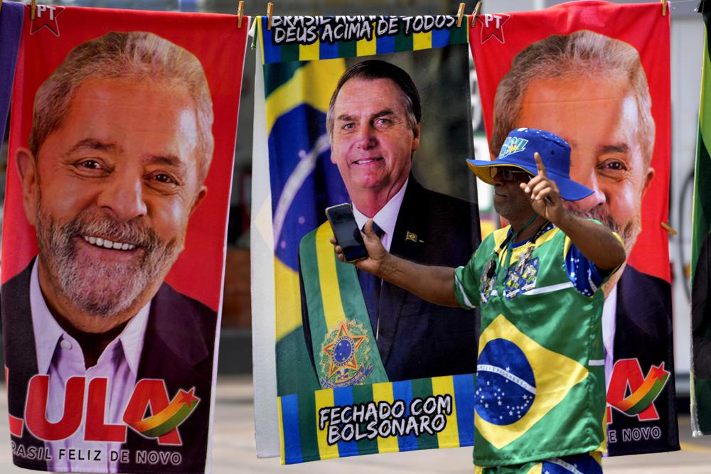 A demonstrator dressed in the colors of the Brazilian flag performs in front of a street vendor’s towels for sale featuring Brazilian presidential candidates, current President Jair Bolsonaro, center, and former President Luiz Inacio Lula da Silva, in Brasilia, Brazil, Tuesday, Sept. 27, 2022. Nearly a dozen candidates are running in Brazil’s presidential election but only two stand a chance of reaching a runoff: former President Luiz Inacio Lula da Silva and incumbent Jair Bolsonaro. (AP Photo/Eraldo Peres)