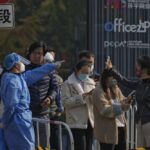 A woman, right, wearing a face mask shows her health check QR code as she and others line up to get their routine COVID-19 throat swabs at a coronavirus testing site in Beijing, Tuesday, Nov. 8, 2022. Police in northeastern China say seven people have been arrested following a clash between residents and authorities enforcing COVID-19 quarantine restrictions. (AP Photo/Andy Wong)