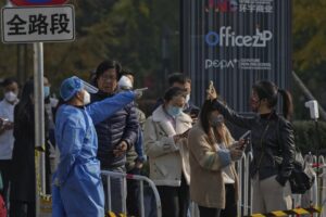 A woman, right, wearing a face mask shows her health check QR code as she and others line up to get their routine COVID-19 throat swabs at a coronavirus testing site in Beijing, Tuesday, Nov. 8, 2022. Police in northeastern China say seven people have been arrested following a clash between residents and authorities enforcing COVID-19 quarantine restrictions. (AP Photo/Andy Wong)