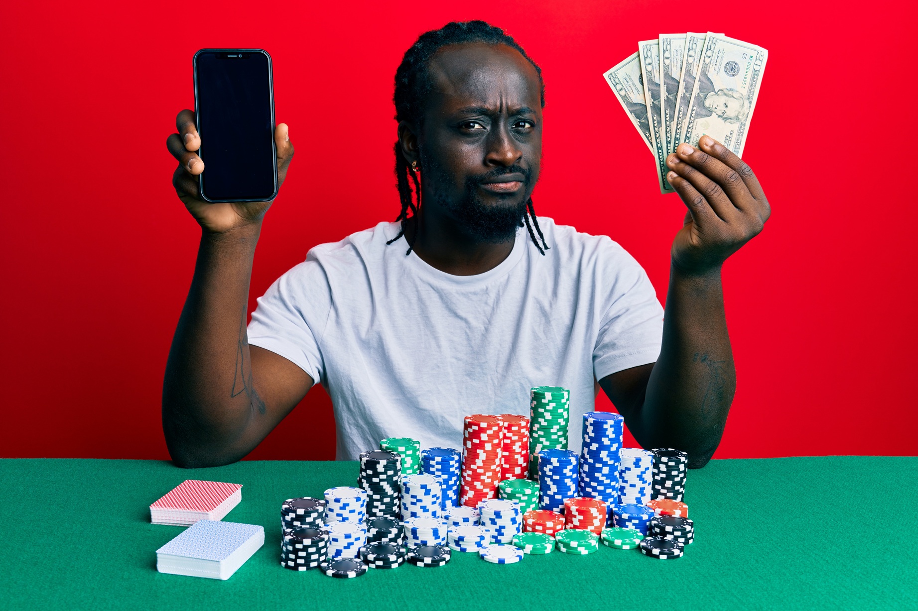 Handsome young black man playing poker holding smartphone and dollars skeptic and nervous, frowning upset because of problem. negative person.
